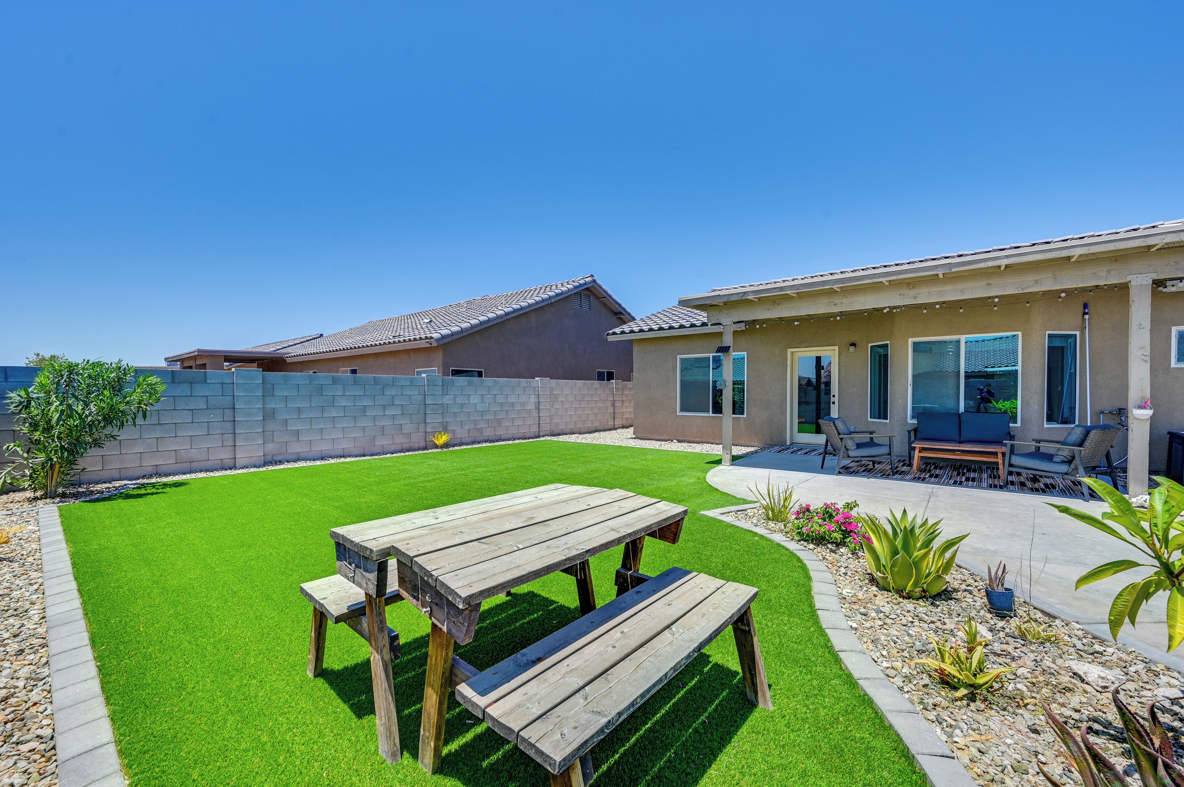 A backyard with a wooden picnic table and bench, a patio with outdoor furniture, and a house with a tiled roof.