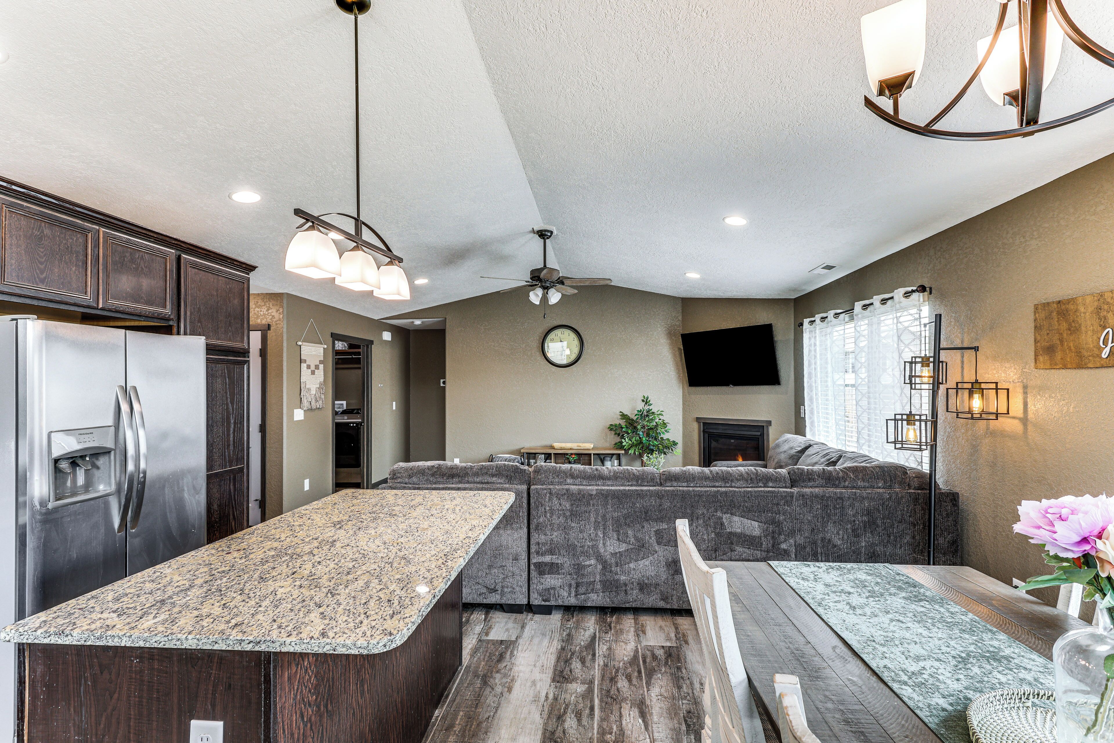 A modern kitchen with dark wood cabinets, a granite countertop, and a built-in island. There is a ceiling fan, recessed lighting, and a large window with curtains.