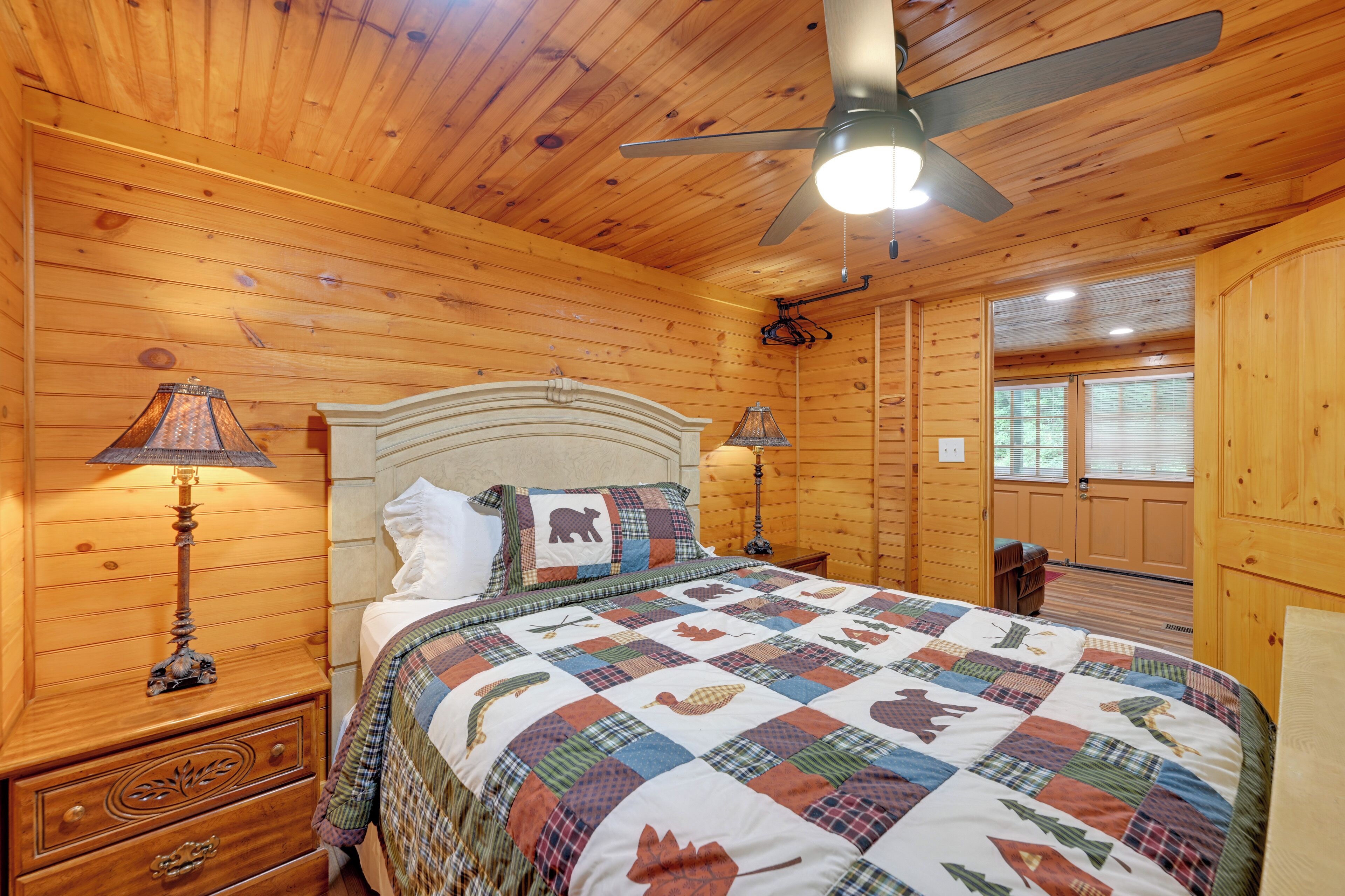 A wooden cabin bedroom with a bed, two bedside lamps, a ceiling fan, and a window.
