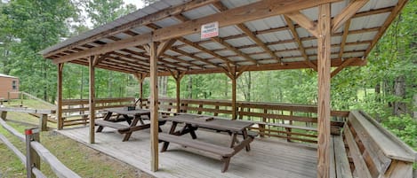 A wooden pavilion with picnic tables, surrounded by a wooden deck and a metal roof, set in a wooded area.