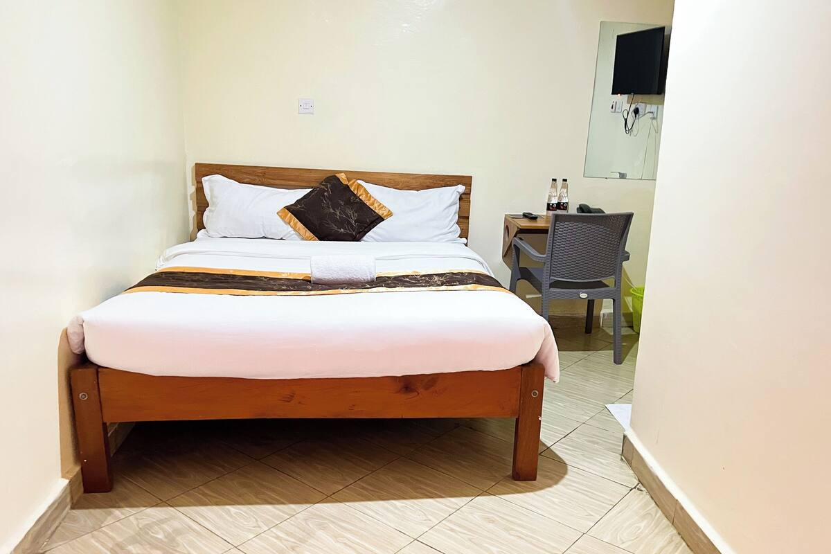A small, neatly arranged bedroom with a single bed, a chair, a desk, and a TV mounted on the wall.