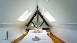 Ein Schlafzimmer mit schrägen Wänden, Dachfenstern, einem Bett mit Kissen, einem Nachttisch, einer Lampe und einem Heizkörper.