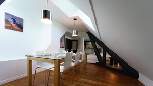 Ein Essbereich mit Glastisch, weißen Stühlen und Pendelleuchten. Eine Treppe mit schwarzen Geländern führt auf eine erhöhte Ebene.