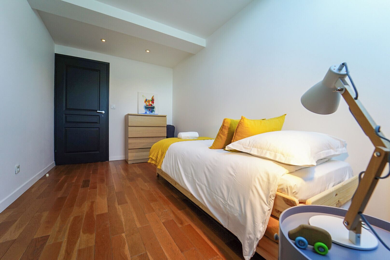 Un dormitorio con piso de madera, una cama con ropa de cama blanca y almohadas amarillas, un escritorio con una lámpara y un aparador.