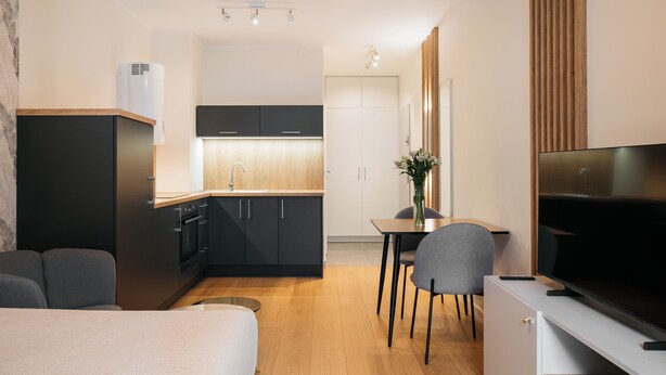 A modern hotel room with a bed, a TV, a dining area with a table and chairs, a kitchen with dark cabinets and a wooden countertop, and a built-in air conditioner. - Native Apartments Barska 69 (Kraków)