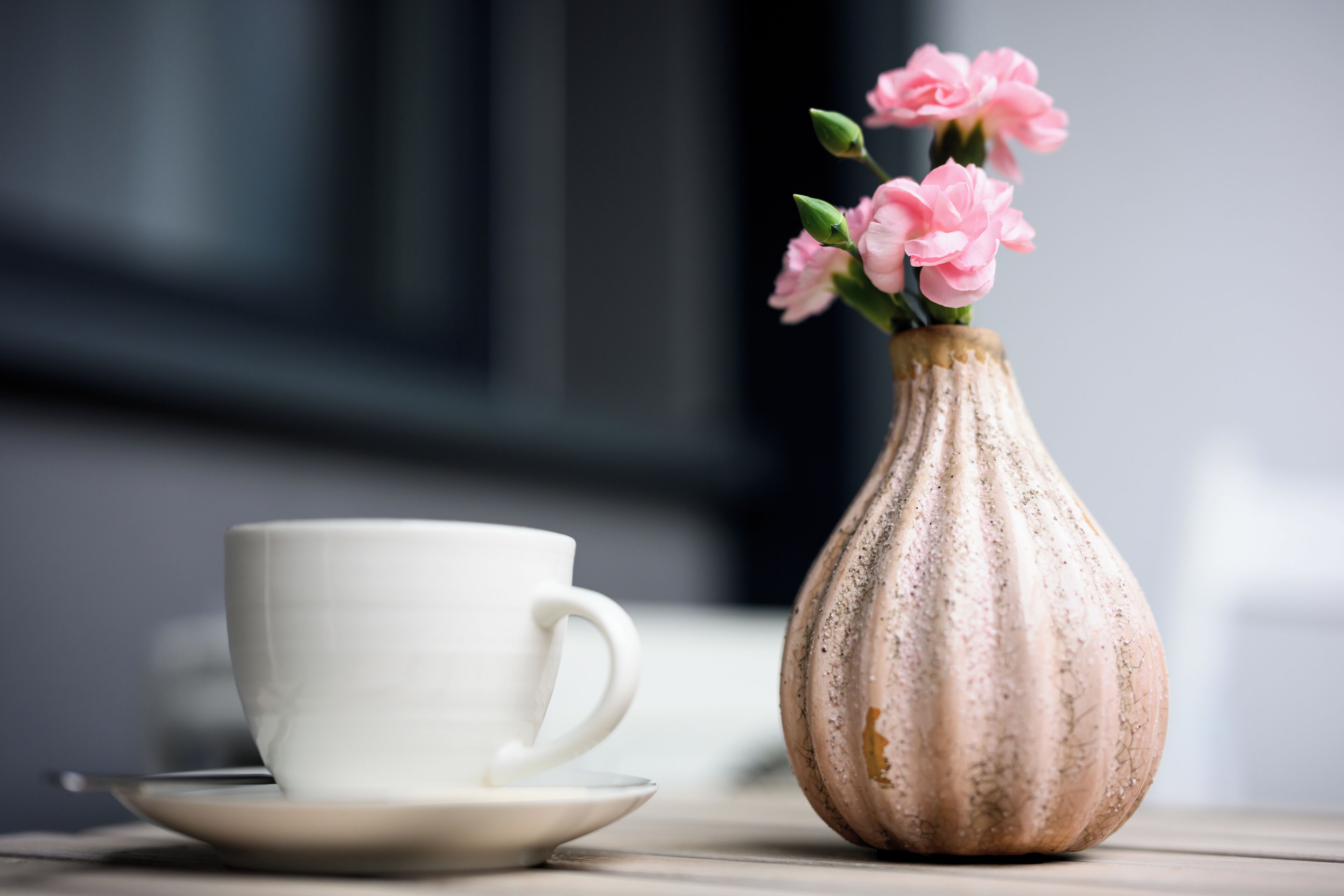 Una taza y un plato blancos sobre una mesa, junto a un jarrón con flores rosadas.