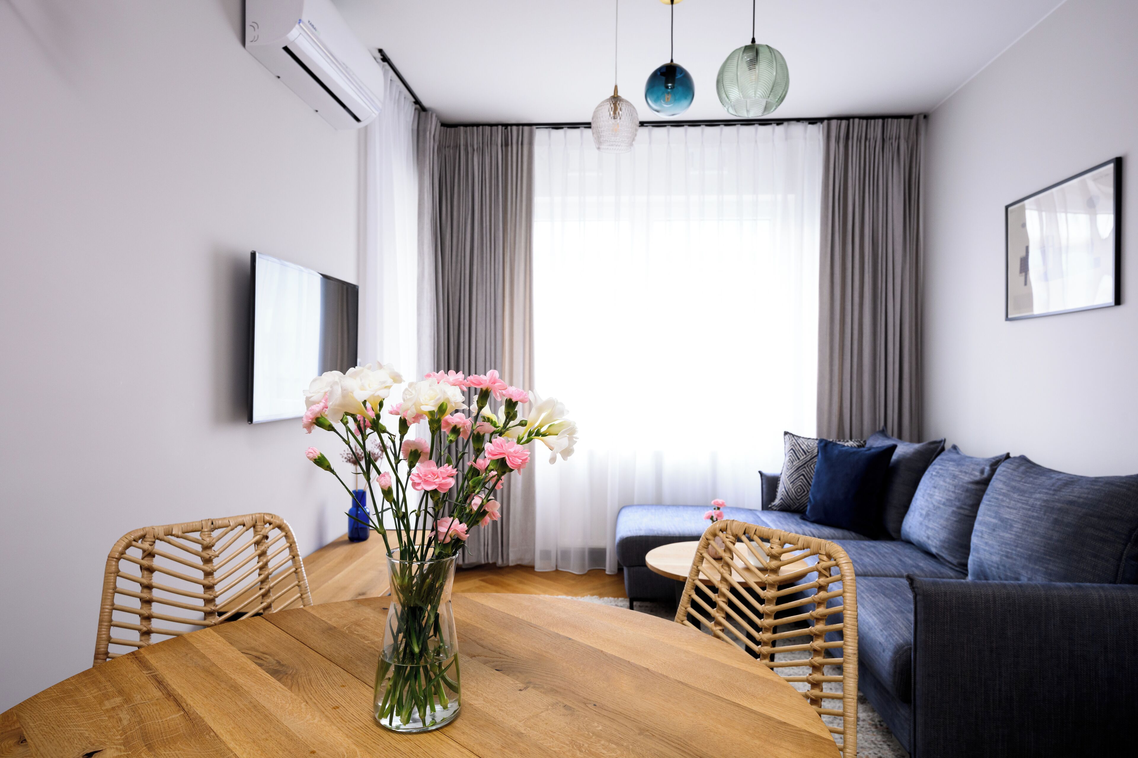 A modern living room with a wooden dining table, wicker chairs, a sofa, and a large window with curtains.