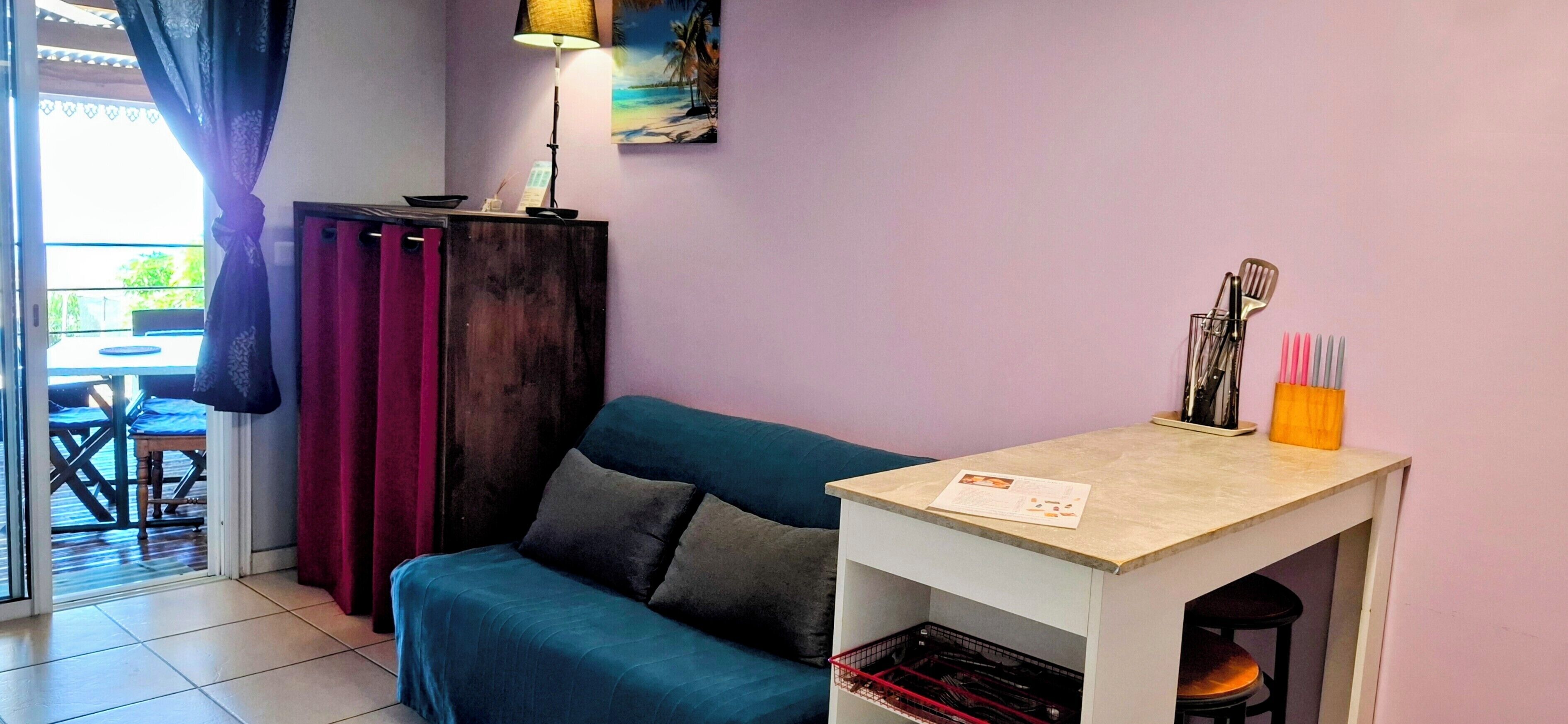 Una habitación con un sofá azul turquesa, una mesita con una lámpara, un cuadro en la pared y vistas al exterior a través de una ventana.