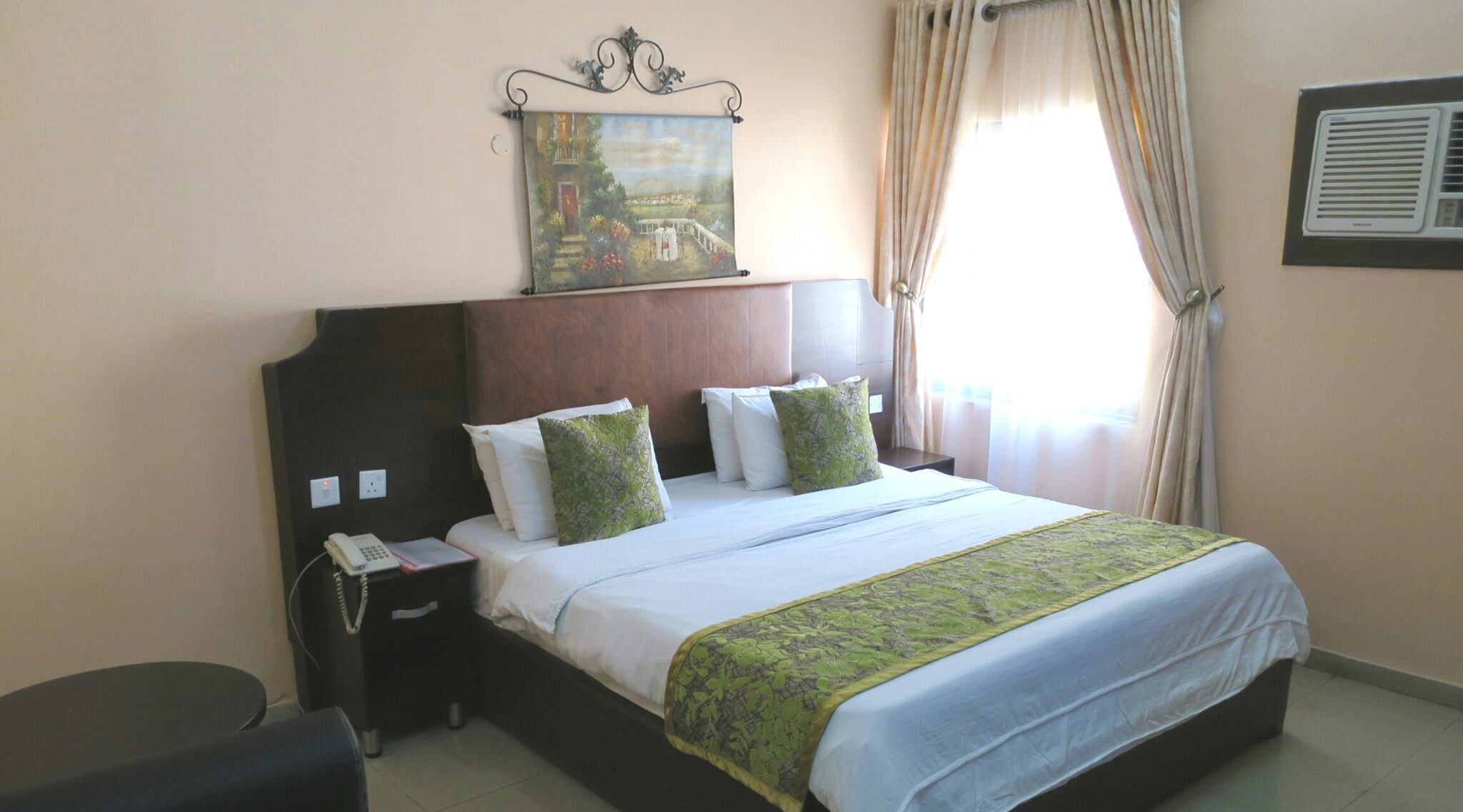 Una habitación de hotel con una cama grande, una mesita de noche con un teléfono, un cuadro sobre la cama y una ventana con cortinas.
