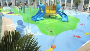 Un espacio de juegos acuáticos con toboganes y fuentes, rodeado de amplios ventanales y plantas de interior.