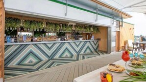 Zona de bar al aire libre con mostrador de diseño en forma de estampada, plantas colgantes y una mesa con comida y bebidas.