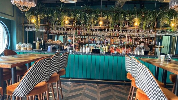 Zona de bar con mostrador verde, diversas opciones de asientos y una variedad de botellas y copas en estantes.