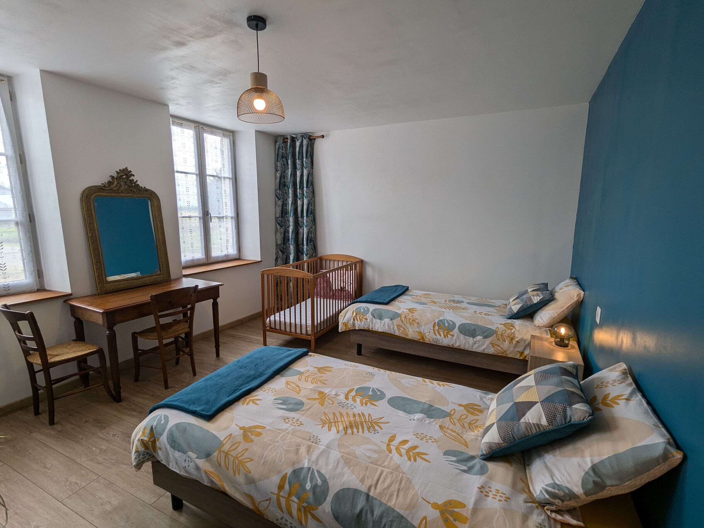 Quarto com duas camas de solteiro, uma escrivaninha de madeira, uma cadeira, um espelho e uma janela com cortinas.