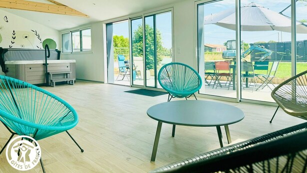 A spacious room with a large sliding glass door leading to an outdoor area, a sofa, a small table, and a chair. - Charming cottage in Vendée with spa and sauna (La Roche-sur-Yon)