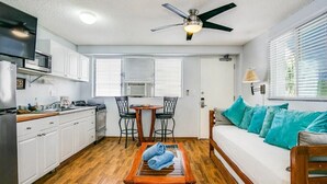 Cocina compacta con gabinetes blancos, un microondas y una estufa. Hay un comedor con una mesa y sillas, y un sofá con cojines color turquesa. Se ve un ventilador de techo, y el cuarto tiene piso de madera.