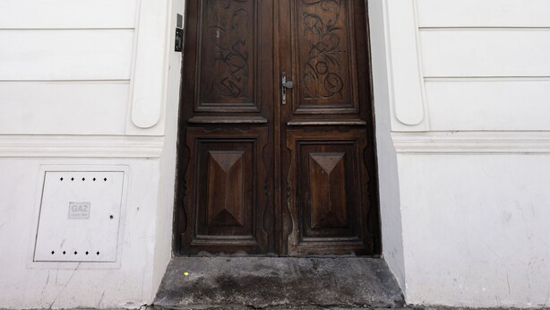 Puerta doble de madera con intrincados grabados, ubicada en la fachada blanca de un edificio con un ventanal arriba.