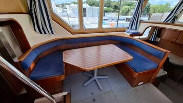 Interior de barco com área de estar curvada, mesa central e janelas com vista para a água e barcos atracados.
