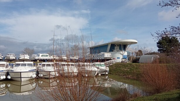 Ein Yachthafen mit mehreren vertäuten Booten, ein großes Gebäude mit markanter, geschwungener Dachform und ein kleiner Wasserbereich, der die Boote reflektiert.