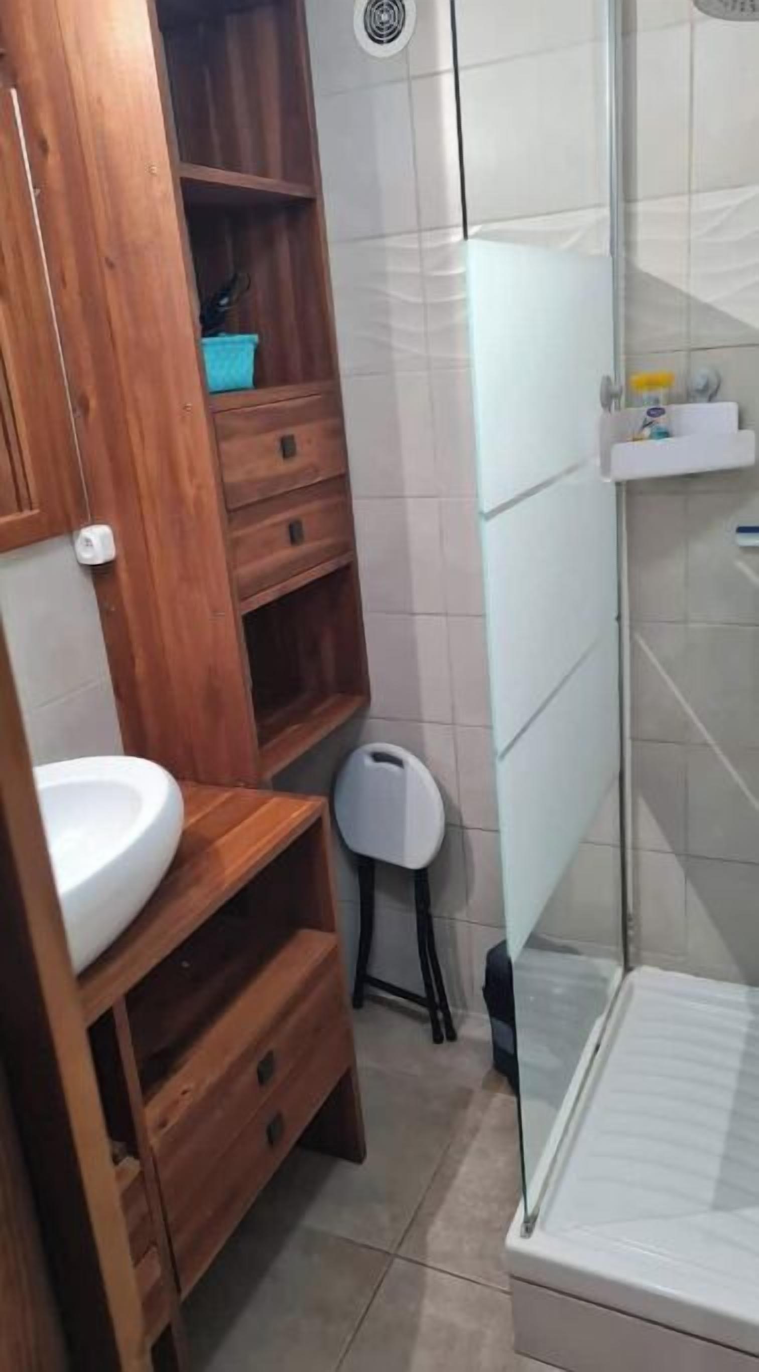 Une salle de bain avec un meuble-lavabo en bois, un lavabo blanc, un petit tabouret et un espace douche.