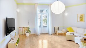 A modern living room with a wooden herringbone floor, a white sofa with yellow pillows, a flat-screen TV mounted on the wall, and a large window with white curtains. - HB apartments Astoria (Tbilisi)