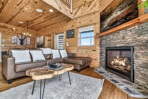 A cozy cabin interior with a stone fireplace, wooden walls, a leather sofa, and a chandelier.