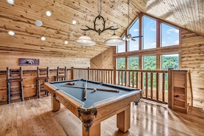 Une table de billard dans une pièce en bois, dotée d’un balcon et de grandes fenêtres.