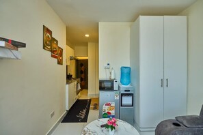 Uma cozinha compacta com geladeira, micro-ondas e dispenser de água. Um tapete preto com a palavra 'Kitchen' escrita nele está no chão.