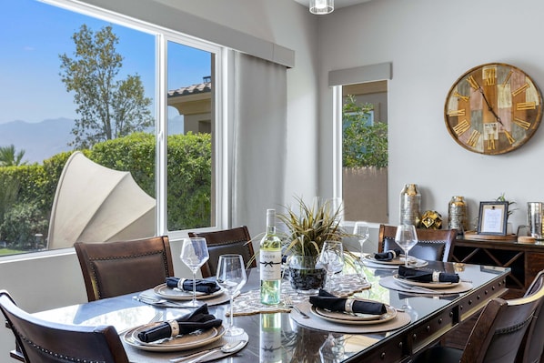 A dining table set with plates, glasses, and a bottle of wine, with a large clock on the wall and a view of greenery outside. - Diamond Palms at Desert Princess Resort (Cathedral City)