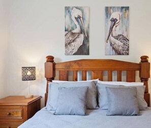 A bedroom with a wooden headboard, a bedside table with a lamp, and two wall-mounted paintings of pelicans.