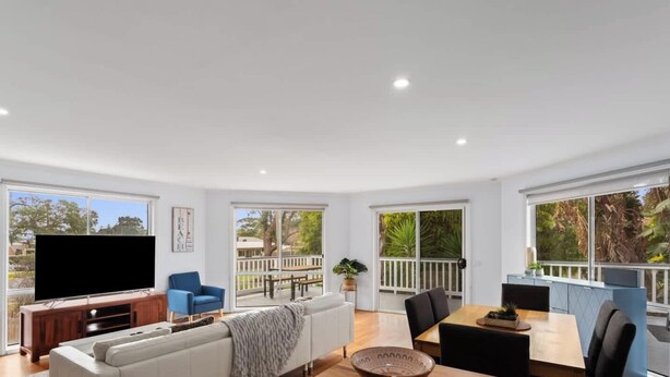 A spacious living area with a flat-screen TV, a sofa, a dining table, and a balcony with a view of the outdoors.