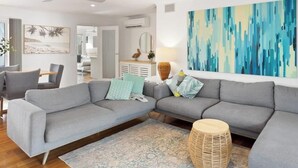 Un salon moderne avec un canapé d’angle gris, un coin repas comprenant une table et des chaises, une grande peinture abstraite et un ventilateur de plafond.