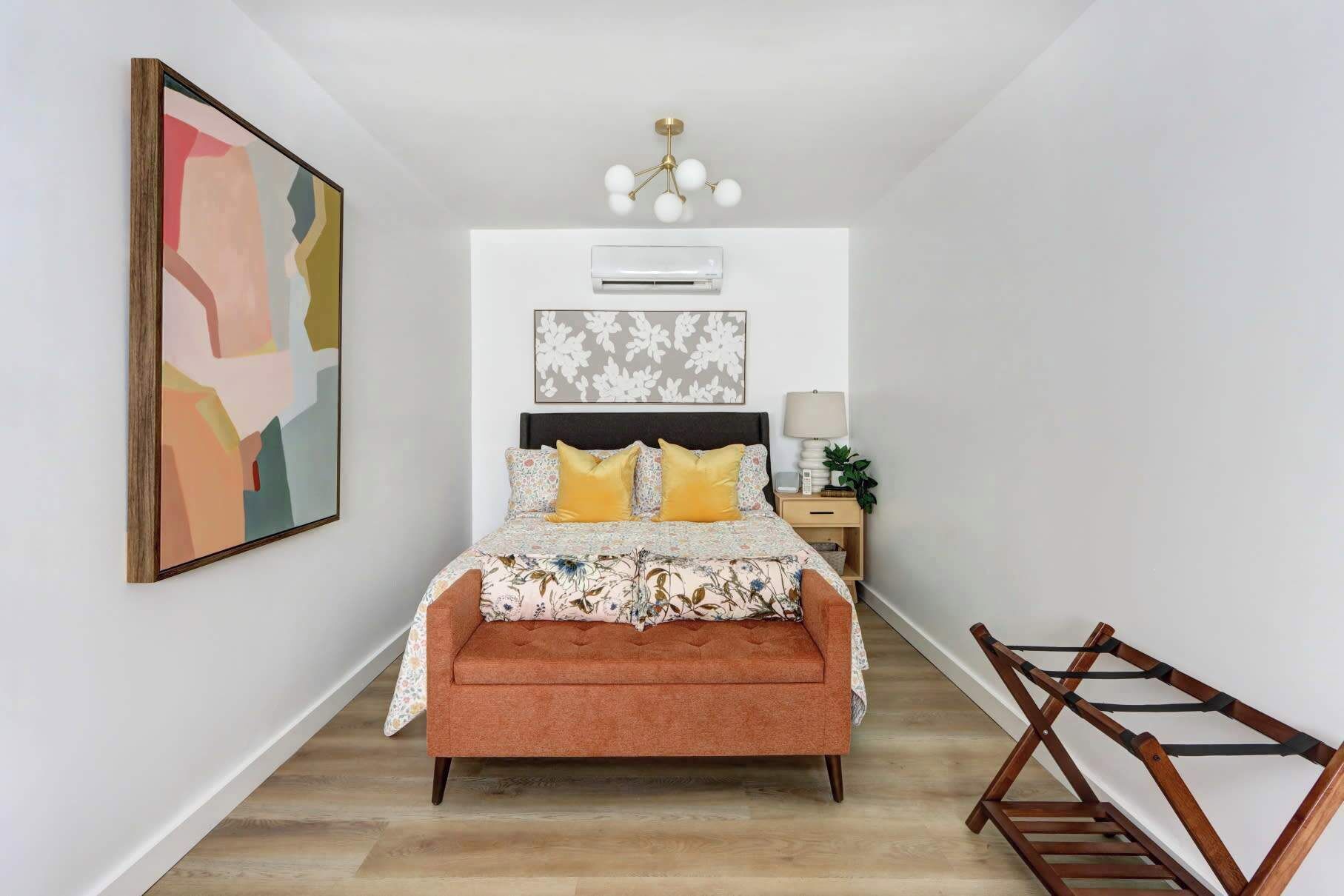 A bedroom with a bed, a nightstand, a lamp, a wall-mounted air conditioner, and a wooden chair.