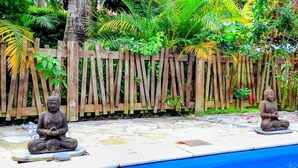 Due statue di Buddha su piattaforme in pietra, vicino a una piscina, con un recinto in legno e vegetazione rigogliosa sullo sfondo.