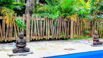 Deux statues de Bouddha sont assises sur des plateformes en pierre, au bord d’une piscine. Une clôture en bois et une végétation luxuriante se trouvent en arrière-plan.