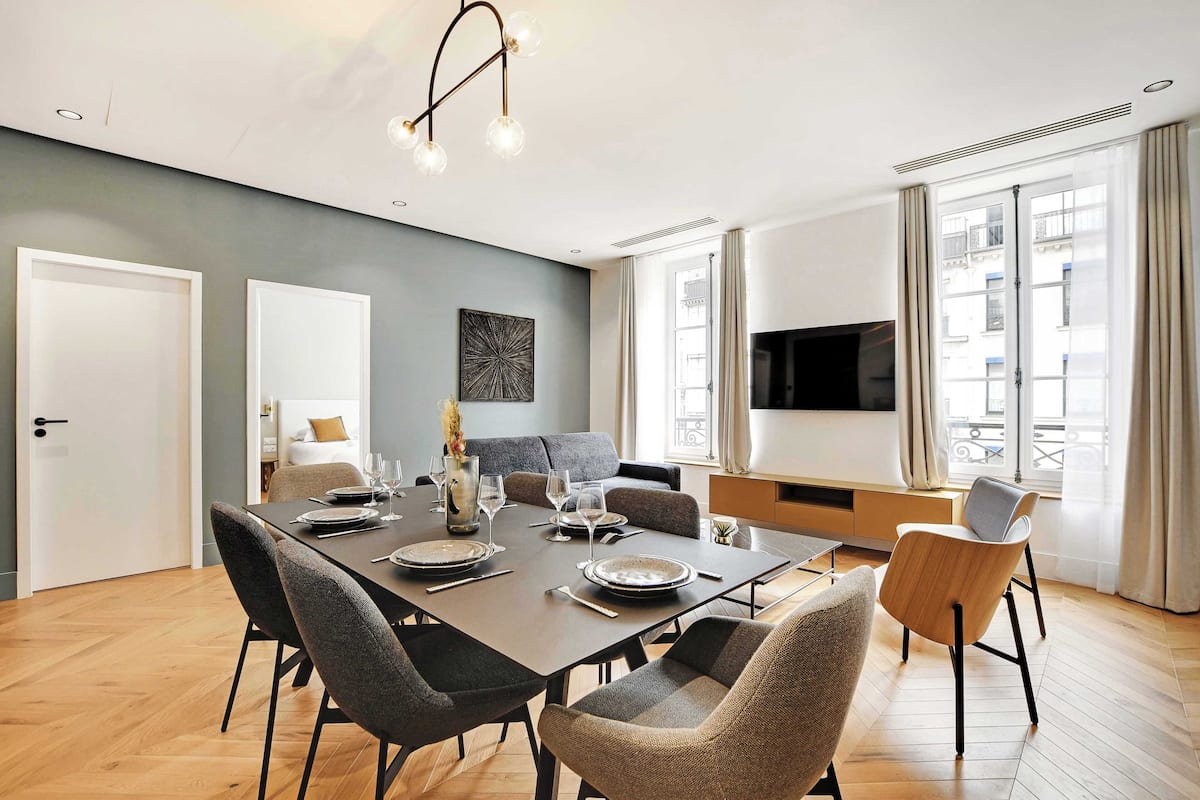 A modern dining area with a large table set for six, surrounded by chairs, a sofa, and a TV.