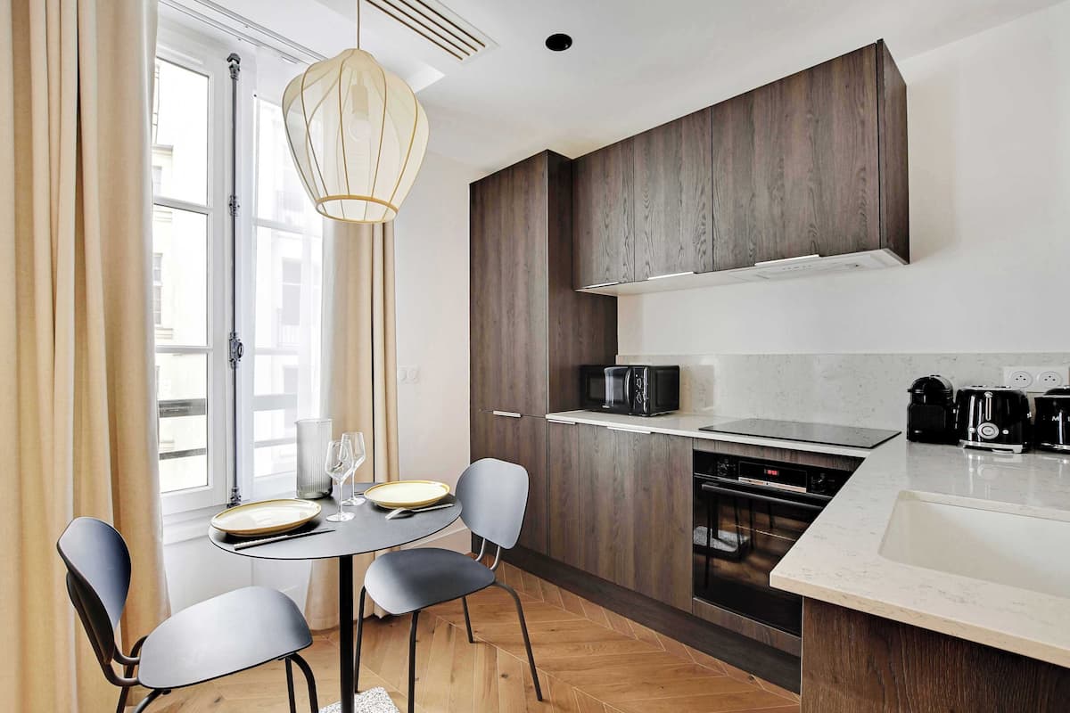 A modern kitchen with a dining area, featuring a large window, wooden cabinets, and a white countertop.