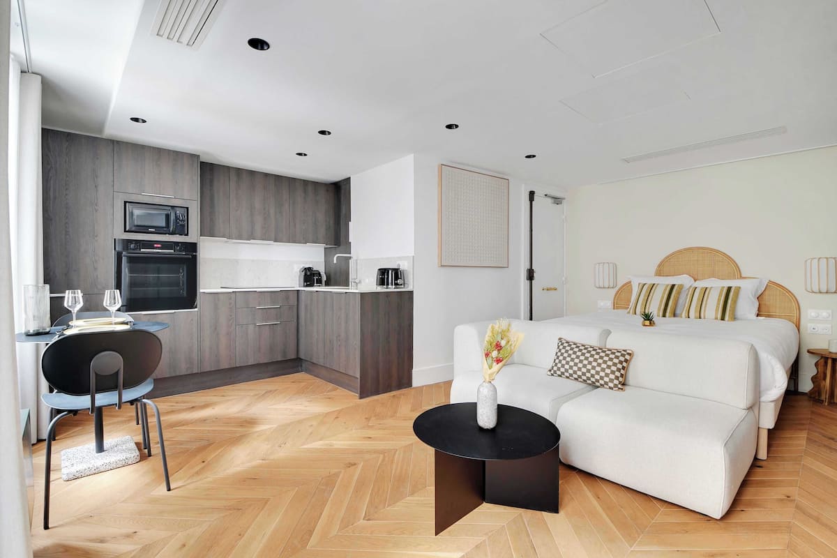 A modern hotel room with a kitchenette, a dining area, and a bed with a checkered pillow.
