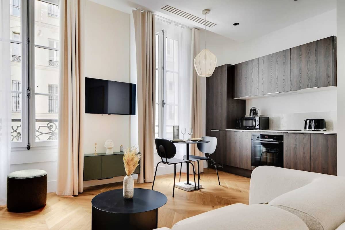 A modern living room with a sofa, a round black coffee table, and a TV mounted on the wall. There is a dining area with a table and chairs, and a kitchen with wooden cabinets and appliances.