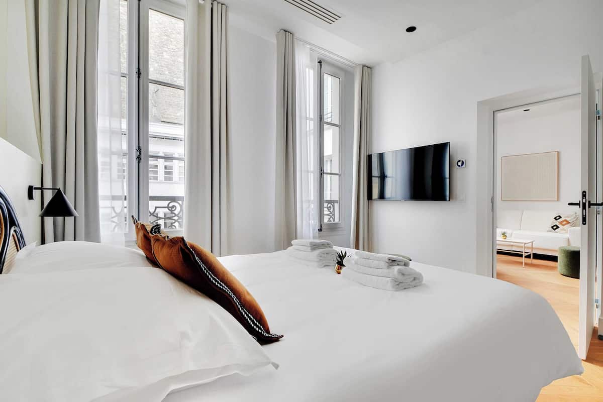 A hotel room with a large bed, a flat-screen TV mounted on the wall, and a view of the city through large windows.