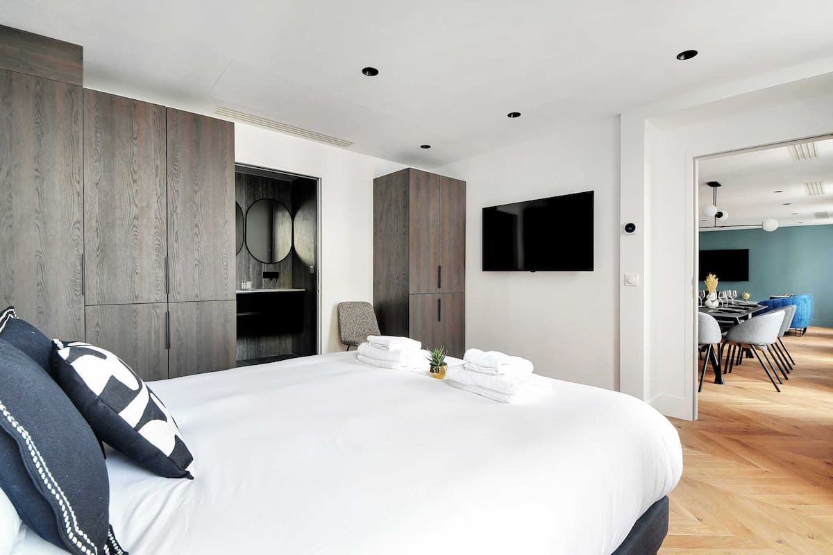 A modern bedroom with a large bed, built-in wardrobes, a flat-screen TV, and a dining area with chairs and a table.