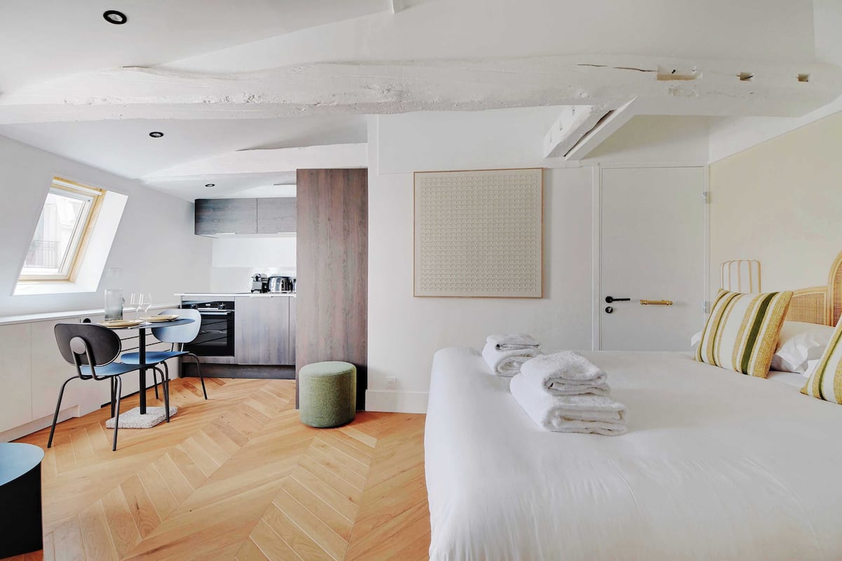 A modern hotel room with a wooden herringbone floor, a bed with white linens, a dining area with a table and chairs, and a kitchenette in the background.