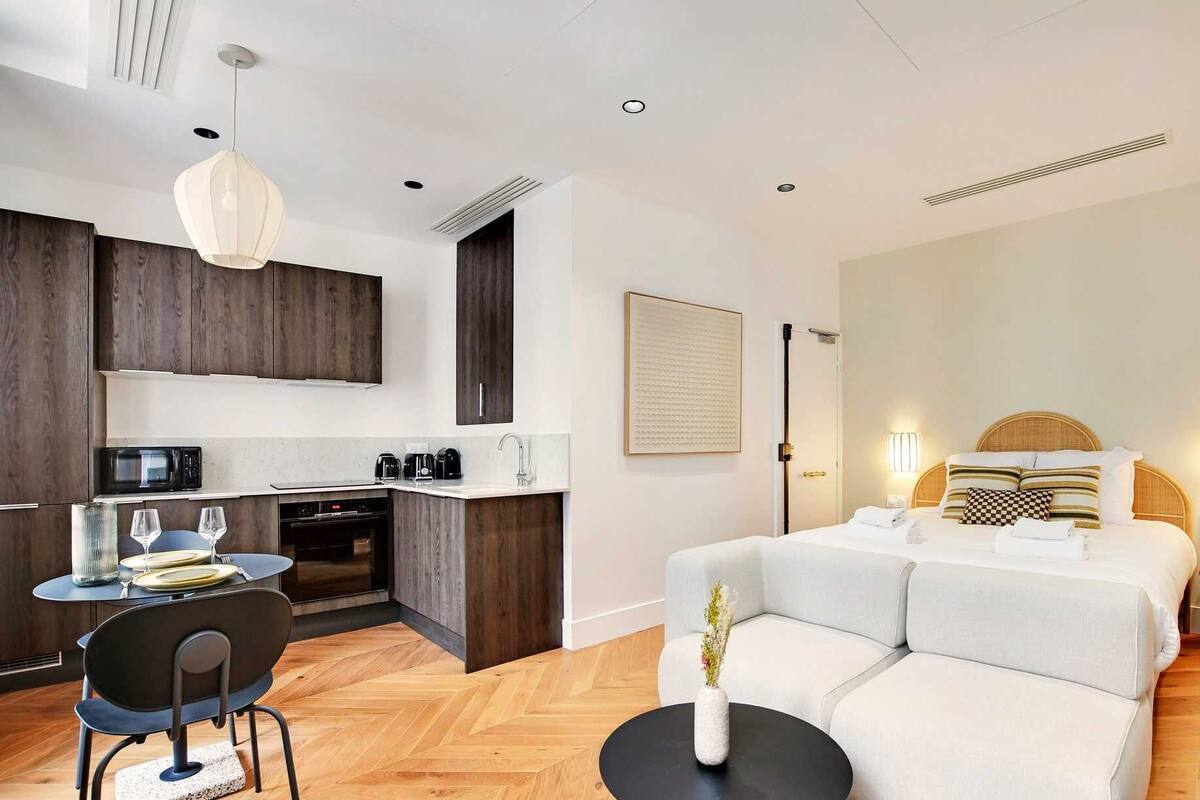 A modern hotel room with a kitchenette, a sofa bed, a dining area, and a large window.