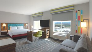 Habitación de hotel con cama, escritorio, silla, televisión y un amplio ventanal con vistas a la ciudad.