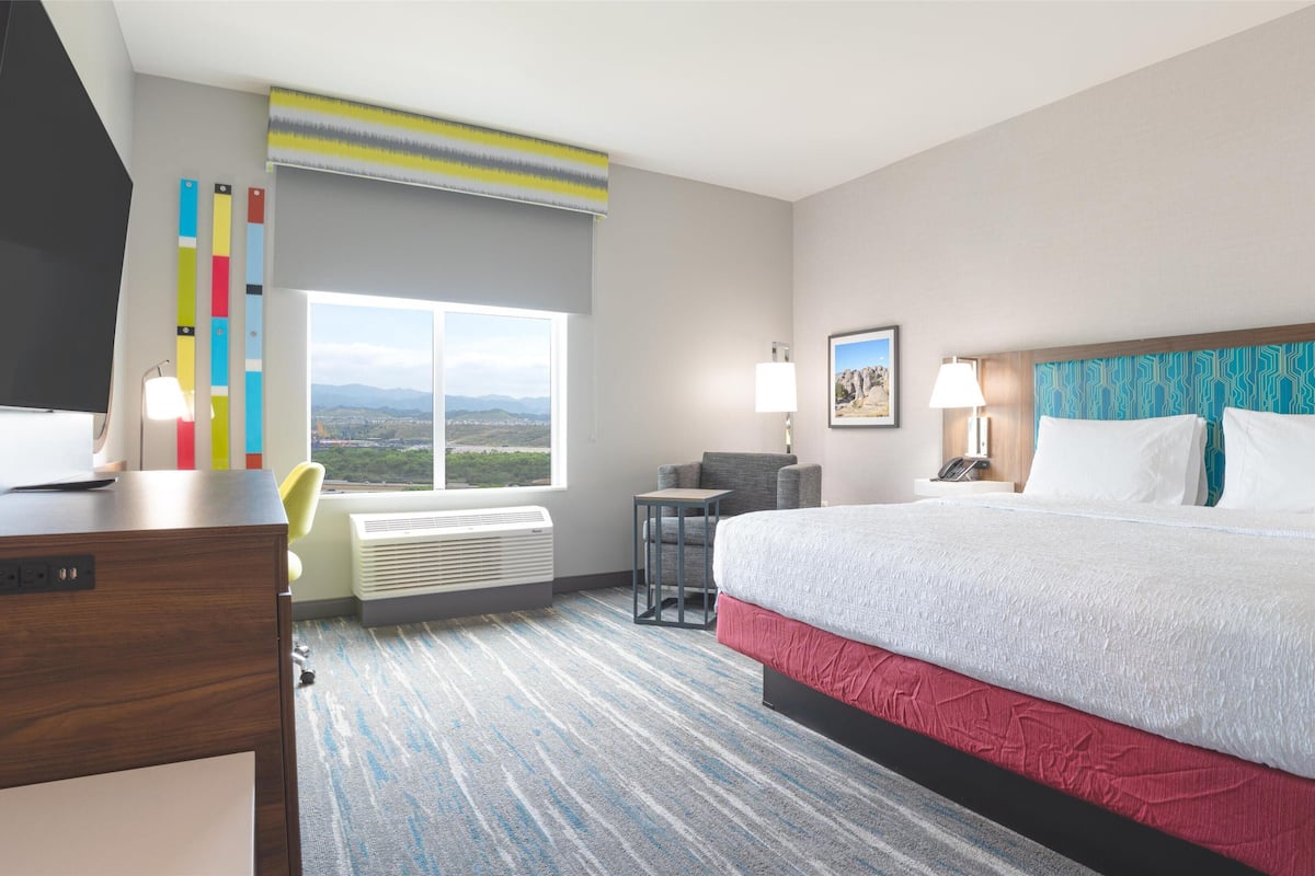 Habitación de hotel con una cama grande, un escritorio con televisión de pantalla plana, una silla, una mesita pequeña, una lámpara y vistas a las montañas desde la ventana.