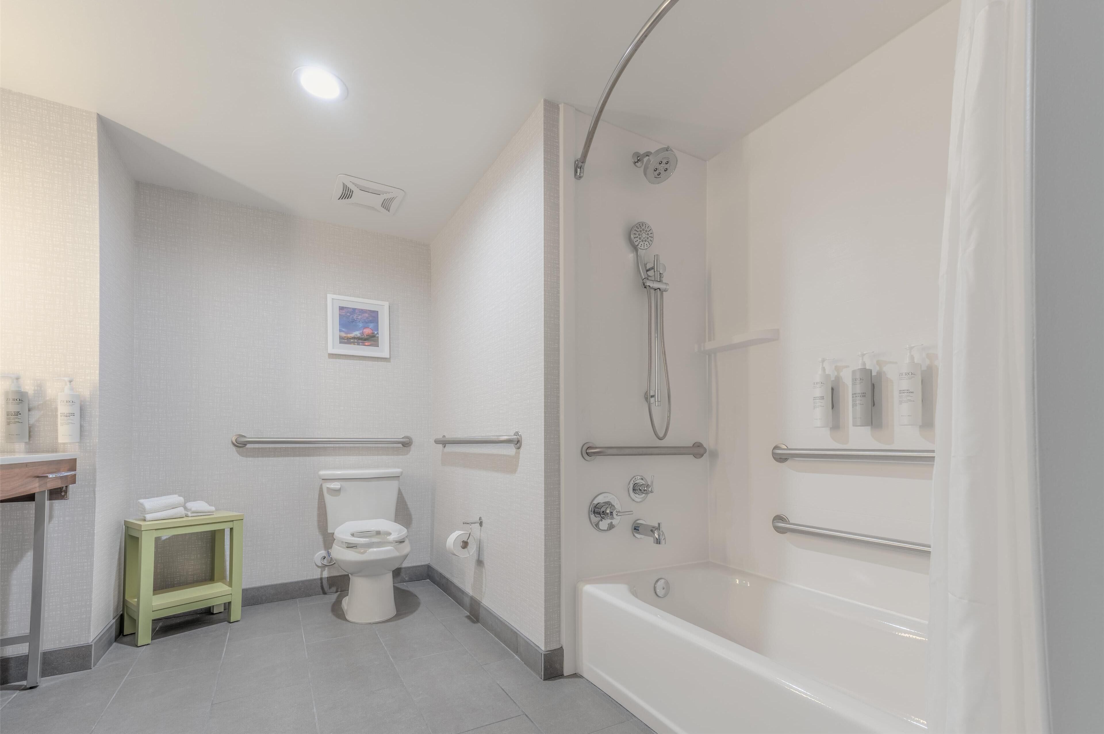 Un baño moderno con inodoro, ducha y bañera. Hay barras de apoyo y un pequeño estante con artículos de aseo.