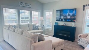 A living room with a beige sectional sofa, a fireplace, a TV mounted on the wall, and a sign that reads "EVERY SUMMER".