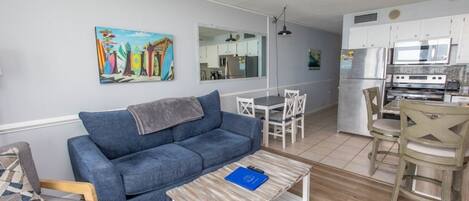 A living room with a blue sofa, a coffee table, and a dining area with white chairs. A kitchen with white cabinets and appliances is visible in the background.