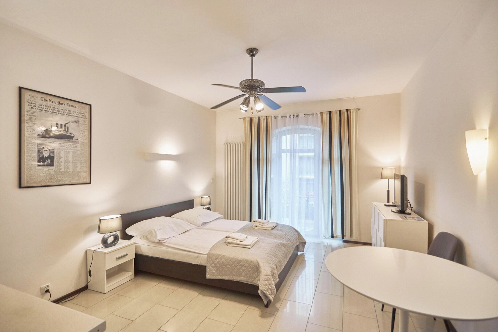 A hotel room with a bed, bedside tables, a desk, a chair, a ceiling fan, and a window with curtains.