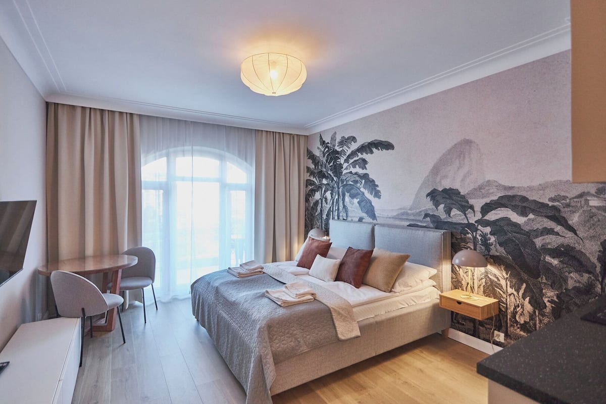 Une chambre moderne avec un grand lit, un bureau et une chaise. On y trouve une fenêtre avec des rideaux, une fresque murale et un luminaire au plafond.