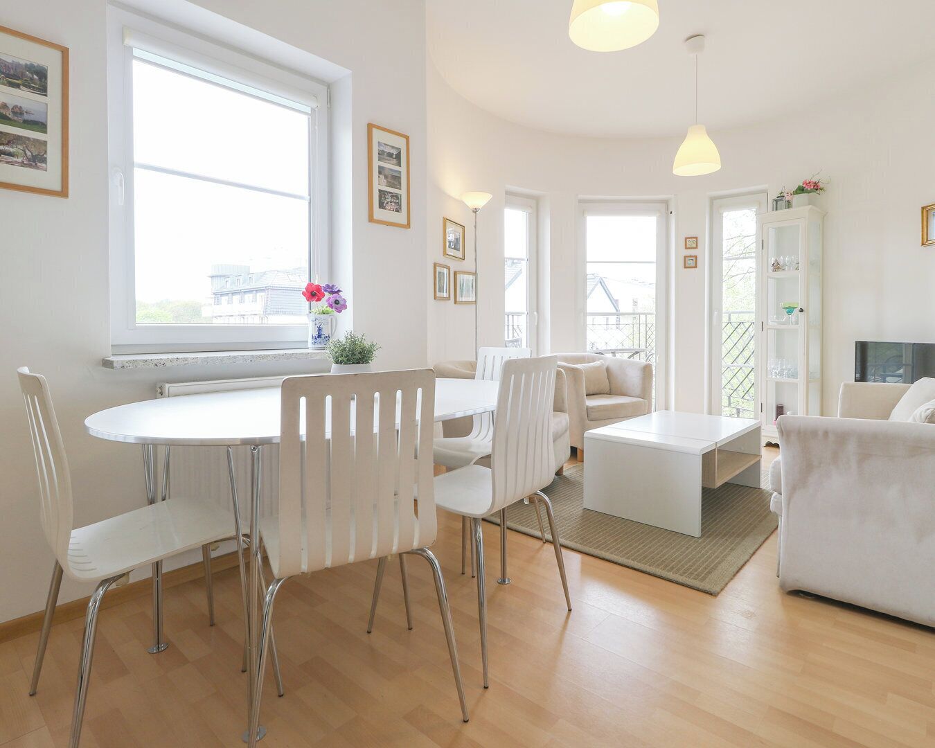 Una sala de estar moderna con comedor, muebles blancos, ventanas amplias y un balcón.